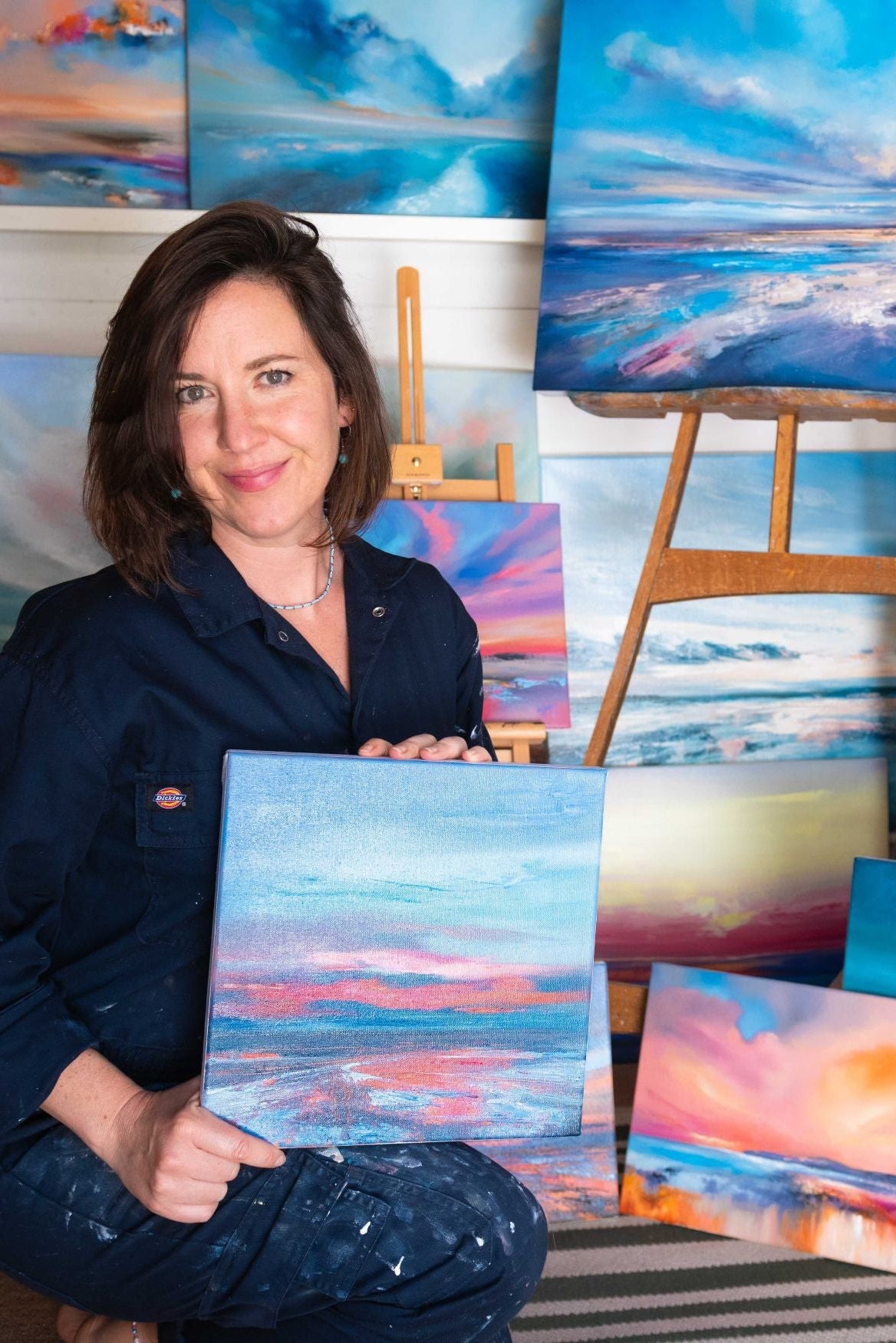 Amy Jobes holding an oil painting with various other artworks displayed around her.